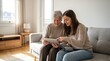 © La Neve - Intergenerational Connection: A young woman warmly engages with an elderly woman on a comfortable sofa, both engrossed in a digital tablet, symbolizing connection and shared moments.