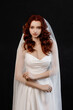 © Kaplitskaya Love - Portrait of a young bride with wavy red hair wearing a white strapless wedding gown and sheer veil, standing against a plain black background in soft studio