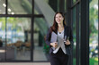 © MINAE - Young Asian businesswoman walking through a bright office hallway, carrying a laptop and coffee, symbolizing a dynamic professional lifestyle and entrepreneurial spirit