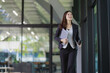 © MINAE - Businesswoman smiling while walking confidently, carrying a laptop and coffee cup in a modern office environment, representing success, productivity, and a professional career path
