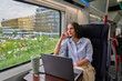 © DusanJelicic - Businesswoman looking out the train window while commuting, reflecting on work and life during a journey, with a laptop and coffee cup ready to work or relax