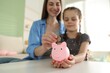 © New Africa - Woman with her daughter putting coin into piggy bank at home, selective focus