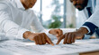 © halcon1 - Two professional male architects lean over a large table and point at architectural blueprints, collaborating and discussing details of a building project in a bright office