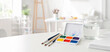 © SerPak - Watercolor palette and pencils arranged on a white table next to a jar of water and paper. Bright minimal art workspace with soft natural light and copy space for text.