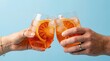© Rafiqul - Closeup of two people clinking glasses of aperol spritz cocktail with orange slices on a light blue background