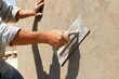 © Studio - Professional builder applying wet plaster to concrete wall with metal trowel at outdoor construction site, close-up view of hands working diligently in bright sunlight.