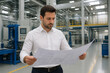 © Anastasiia - A man in a white shirt is reviewing blueprints inside a large industrial factory setting.