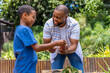 © wavebreak3 - African American father and teenage son leaning over raised wood planter holding soil and seedling