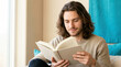 © Len_mik - Young man with long hair reading a book at home. Smiling male reader enjoying a novel indoors. Leisure and relaxation concept