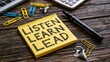 © Behram - Sticky note featuring listen learn lead text sits on wooden table with a pen and various paper clips set.