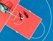 © 晨啸 周 - aerial view of two students playing one-on-one basketball on a vibrant red and blue school court. Bright sunlight casts long dramatic shadows, highlighting active outdoor sports and fitness.