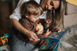 © qunica.com - A mother reads a colorful board book to her toddler as they sit on the floor. The parent and child share a quiet moment exploring pictures, toys and words together.