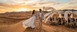 © Maxime - Mariés marchant main dans la main dans les dunes de sable du désert au coucher du soleil