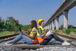 © Verin - two happy diverse female civil engineers wearing hard hat and safety vest sitting back to back on railway tracks,an african woman working on laptop and talking together