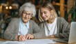© Selvi - An elderly woman with white hair and glasses writes with a pen while a younger woman watches beside a laptop at a wooden table
