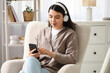 © Pixel-Shot - Pretty young woman with mobile phone and headphones listening to audiobook on armchair in living room