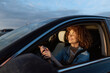 © SHOTPRIME STUDIO - Cheerful woman driving a car, smiling with bright eyes, sunlit moment through the window, relaxed posture, denim jacket, striped top, natural colors, scenic road in the background