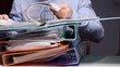 © Aliaksandr - Person arranging and organizing documents within several full binders stacked on a desk, representing a high volume of administrative office work and bureaucracy