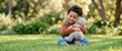 © Xenia - Happy toddler boy hugging a plush teddy bear while sitting on green grass. Smiling child in denim overalls playing in a sunny park. Wide banner with copy space