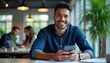 © miss irine - Young Hispanic man in blue shirt uses mobile phone at modern office desk. He smiles positively at camera, works with colleagues nearby. Bright workplace with plants and lights.