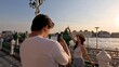 © blueringmedia - Man Taking Smartphone Photo of Woman at Riverside Pier During Sunset