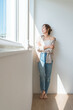 © buritora - A woman (model, fashion) standing by a window bathed in sunlight.