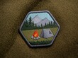 © HooSv - Hyperrealistic sharp focus macro photo of a hexagonal embroidered patch showing a campsite with a tent, campfire, mountains, and pine trees.