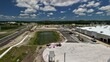 © AmazingAerialAgency - Aerial view of a construction site featuring a large warehouse building, a retention pond, heavy machinery, and a paved road under a blue sky in the United States.