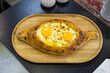 © FrenchDailyLife - Freshly baked Adjaruli khachapuri with cheese and two runny egg yolks rests on a rustic wooden board. This traditional Georgian pastry represents authentic Caucasian comfort food and gourmet dining cu