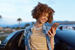 © SHOTPRIME STUDIO - Young woman with curly hair smiles while checking her phone beside a car on a sunny day