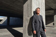 © f_bossa - Man in charcoal suit leans against a textured concrete pillar within a brutalist structure during bright daylight with sharp shadow play across the pavement.