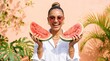 © Mitali Creative  - Smiling woman with hair in a bun wearing pink sunglasses and a white shirt holding two large slices of fresh red watermelon against a soft peach wall with green tropical palm leaves