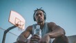 © ЮРИЙ ПОЗДНИКОВ - Resting on a basketball court, a dark-skinned African American man in his 20s wears a grey tank top and headphones while holding a bottle of water.