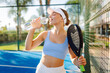 © maxbelchenko - Young woman in sportswear, holding racket, drinks water after game of padel tennis. Athletic woman holds racket while standing on blue court. Concept of relaxation, fun, and active lifestyle.