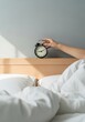 © Ariestia - Woman's Hand Reaching to Turn Off Vintage Alarm Clock on Wooden Headboard in Bright Minimalist Bedroom