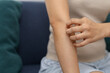 © Ratirat - Frustrated woman tries to scratch her itchy arm suffering from severe skin discomfort and dry allergy rash using her hand while sitting on couch at home feeling physical irritation and pain