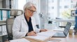 © Eve - Professional senior female doctor or mature physician with glasses examining medical records papers and clinical documentation at office desk with laptop computer in private clinic