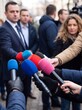 © Shozi - Close-up of multiple colorful microphones held by reporters towards a suited man being interviewed on a city street with people in the background.