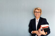 © Westend61 - Portrait of mature businesswoman standing at a wall holding stack of books