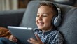 © geniuskp - There is a young boy sitting on a couch with headphones on and looking at a tablet