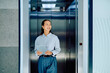 © we.bond.creations - Young Asian businesswoman smiling, holding a digital tablet, and stepping out of a modern elevator in an office building