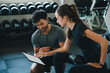 © chokniti - Personal trainer gym woman man clipboard workout consultation fitness coaching exercise, focused adult pair reviewing training plan near dumbbell rack on bench