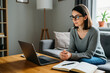 © miss irine - Young woman wears glasses reading laptop screen sitting on sofa with open notebook. Represents everyday cognitive labor, invisible mental workload, gentle productivity, self-directed learning.