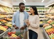 © KIMOG - Happy African American Man Looking at Asian Woman Holding Notebook While Grocery Shopping