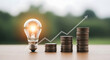 © Aklima - A lightbulb with glowing idea next to rising stacked coins and growth graph outside on table