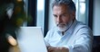 © tiger - mature man with blurred face working intently on a laptop at a windowed office desk in soft natural light, wearing a white shirt with a blurred plant in the foreground