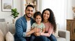 © Nur - A joyful mixed-race family with a young daughter smiling warmly while posing together on a couch in their cozy home living room.