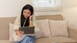 © itchaznong - Asian woman with headphones using digital tablet on sofa at home