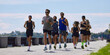 © master1305 - Mixed running club jogging on sunny riverside path with city skyline. Concept of marathon preparation, group fitness, wellness programs and active weekend lifestyle.
