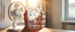 © itlada - Cooling down in hot weather, a hand holds a glass of ice water with condensation, next to a fan and thermometer showing high temperature, representing relief and comfort